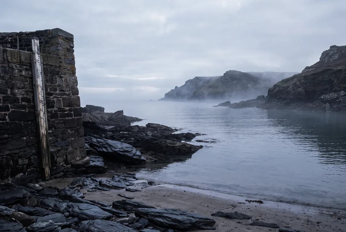 A small cove at low tide with a painted tide staff on the harbor wall and still grey water.