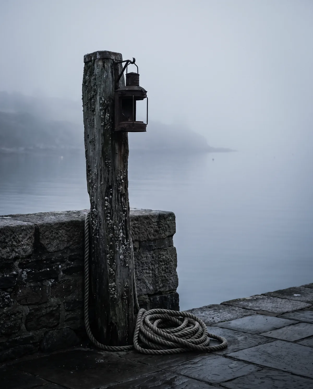 A wooden mooring post with a small lantern at a harbor wall, mist over the water behind it.